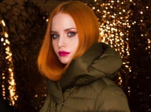 A red haired woman in an olive puffer coat against a backdrop of gold lights
