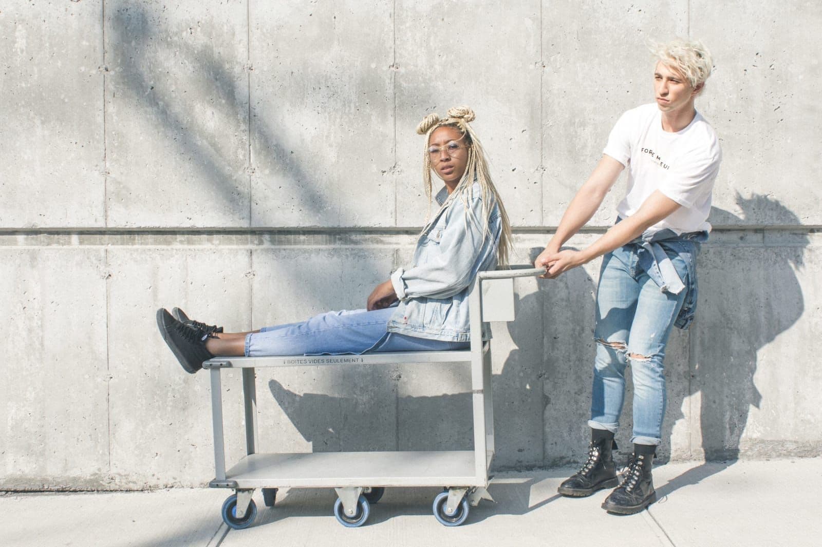 Two people in denim posing beside a metal trolley against a concrete wall
